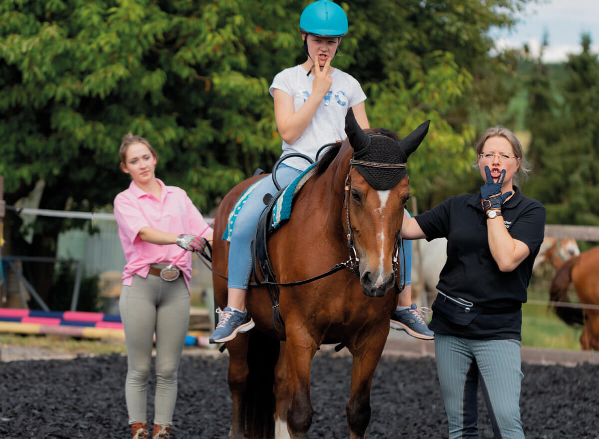 Mädchen sitzt auf einem Pferd während der Therapiestunde mit 2 Therapeuten